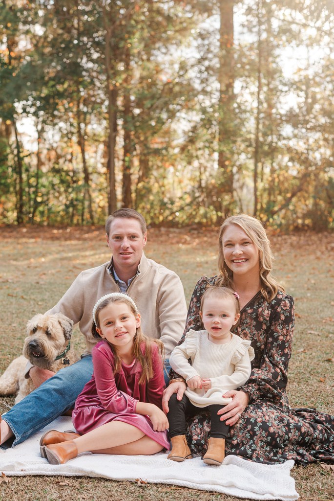 A family of four sitting together on a blanket outdoors, with a dog lying beside them. The adults are casually dressed, while the children are wearing cute outfits. The background features trees with autumn foliage.